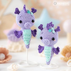 May include: Two handmade crochet seahorse figurines. The seahorses are primarily lavender with purple accents and a mint green belly. They have black eyes and small decorative flowers on their heads. The seahorses are displayed on white stands.