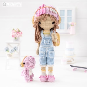 May include: A crocheted doll wearing a pink and white hat, blue overalls, and yellow shoes. The doll is holding a yellow bag. A pink crocheted dinosaur is standing next to the doll.