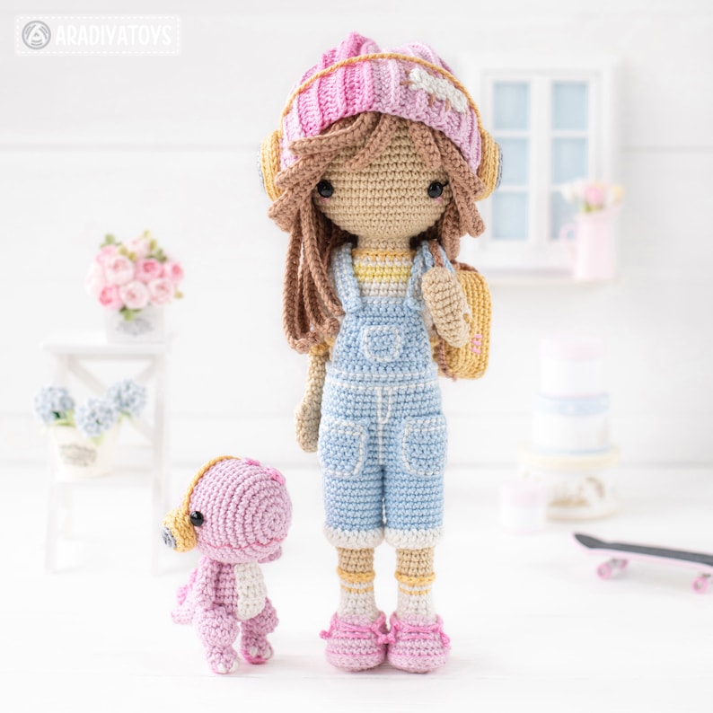 May include: A crocheted doll wearing a pink and white hat, blue overalls, and yellow shoes. The doll is holding a yellow bag. A pink crocheted dinosaur is standing next to the doll.