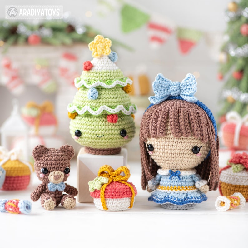 Op de afbeelding: Een verzameling handgemaakte gehaakte kerstversieringen en poppen. Bevat een groene kerstboom met een gele ster, een bruine teddybeer, een pop met een blauwe strik en een geschenkdoos. De versieringen zijn in verschillende kleuren en ontwerpen.