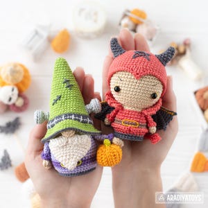 May include: Two small crochet dolls featuring a green gnome with a tall pointed hat and a red devil with horns and wings. The gnome has a white beard and holds a small orange pumpkin. Both figures are crafted with detailed stitching and black eyes.