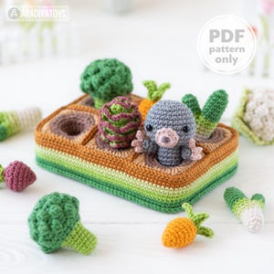 Pode incluir: Um jardim de brinquedo de crochê com uma toupeira cinzenta, brócolis, cenouras e outros vegetais. O jardim é um retângulo marrom, verde e laranja com buracos para os vegetais. O texto "PDF pattern only" está em um círculo branco.