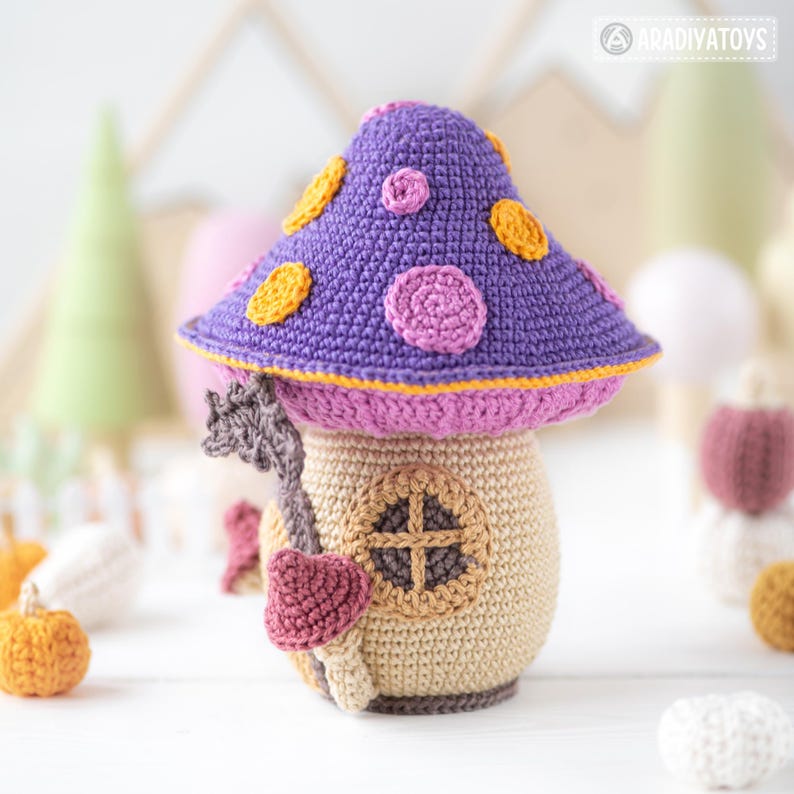 May include: A crocheted mushroom house with a purple cap adorned with yellow and pink circles. The house has a tan base with a small window and a pink door. A whimsical, handmade decorative item.