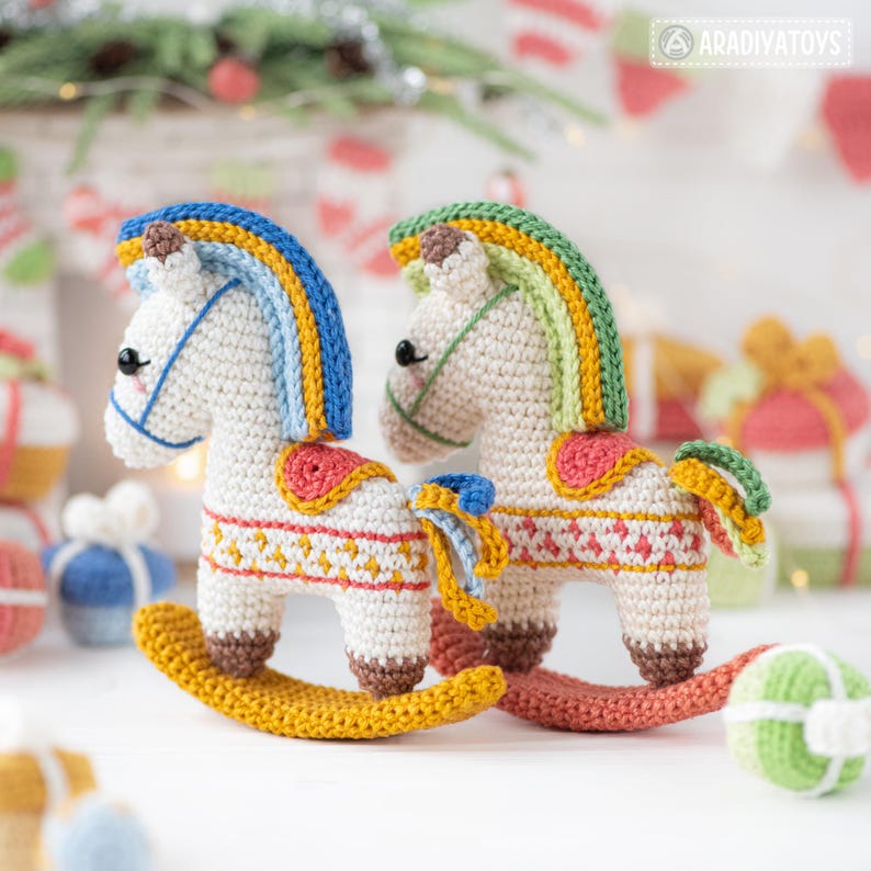 May include: Two handmade crochet rocking horses. The horses are white with colorful manes, saddles, and decorative patterns. The rocking horses have yellow rockers and are decorated with red, blue, yellow, and green accents. The toys are likely for children.