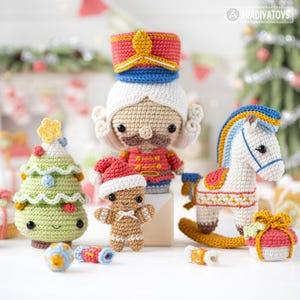Op de afbeelding: Een verzameling handgemaakte gehaakte kerstversieringen, waaronder een notenkraker, kerstboom, peperkoekmannetje, schommelpaard en ingepakt cadeau. De ornamenten zijn in verschillende kleuren, waaronder rood, groen, wit en goud. Het Aradiyatoy's logo is zichtbaar.