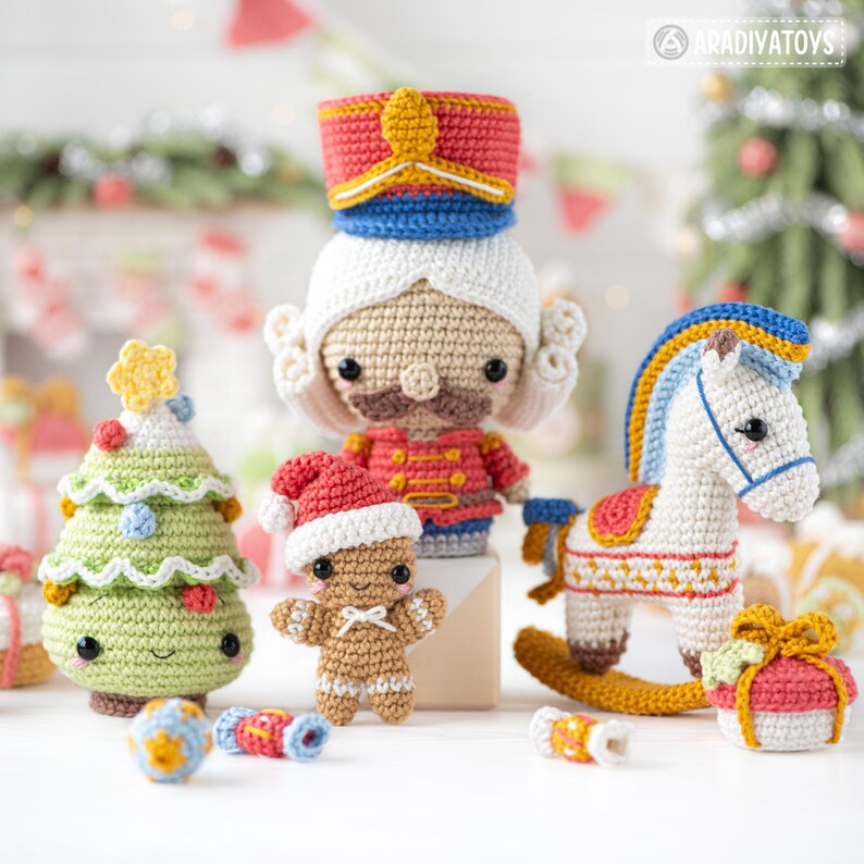 Op de afbeelding: Een verzameling handgemaakte gehaakte kerstversieringen, waaronder een notenkraker, kerstboom, peperkoekmannetje, schommelpaard en ingepakt cadeau. De ornamenten zijn in verschillende kleuren, waaronder rood, groen, wit en goud. Het Aradiyatoy's logo is zichtbaar.