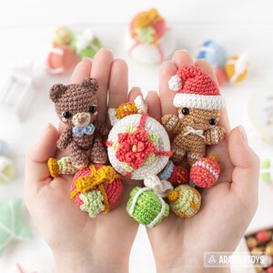 Op de afbeelding: Een verzameling handgemaakte gehaakte kerstversieringen en beeldjes, waaronder een teddybeer met een blauwe strik, een peperkoeken man met een kerstmuts en diverse cadeauvormige ornamenten. De ornamenten zijn in bruin, rood, groen en wit.