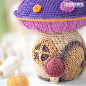 May include: A handmade crochet mushroom house in shades of purple, pink, tan, and brown. The mushroom cap is purple with yellow accents and pink trim. The house features a round window and a small, round, red-purple mushroom. The overall theme is whimsical and decorative.
