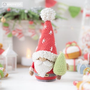 K&ouml;nnte beinhalten: Eine handgefertigte H&auml;kel-Weihnachtsmannfigur mit roter M&uuml;tze, verziert mit wei&szlig;en Schneeflocken und einem flauschigen wei&szlig;en Bommel. Der Weihnachtsmann hat einen wei&szlig;en Bart, einen grauen G&uuml;rtel und h&auml;lt einen kleinen gr&uuml;nen Weihnachtsbaum. Eine festliche Weihnachtsdekoration.