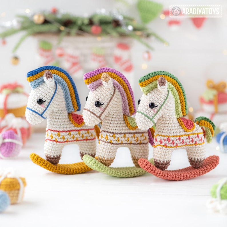 May include: Three handmade crochet rocking horses in a row. Each horse has a white body, colorful mane, and a decorative saddle. The rockers are in shades of yellow, green, and red. The background includes Christmas decorations.