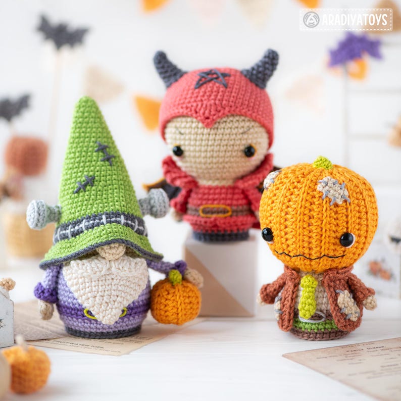 May include: Three crocheted dolls featuring a green gnome with a pointed hat and bolts, a red devil figure with horns and a pentagram, and an orange pumpkin-headed character with a stitched jacket, all crafted with detailed yarn textures.