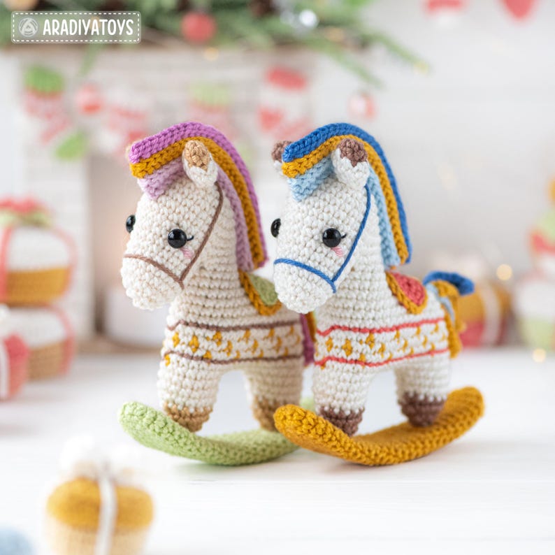 May include: Two handmade crochet rocking horses. The horses are white with colorful manes and saddles. One has a purple, yellow, and orange mane, and the other has a blue, yellow, and brown mane. The rocking horses are on green and yellow rockers.