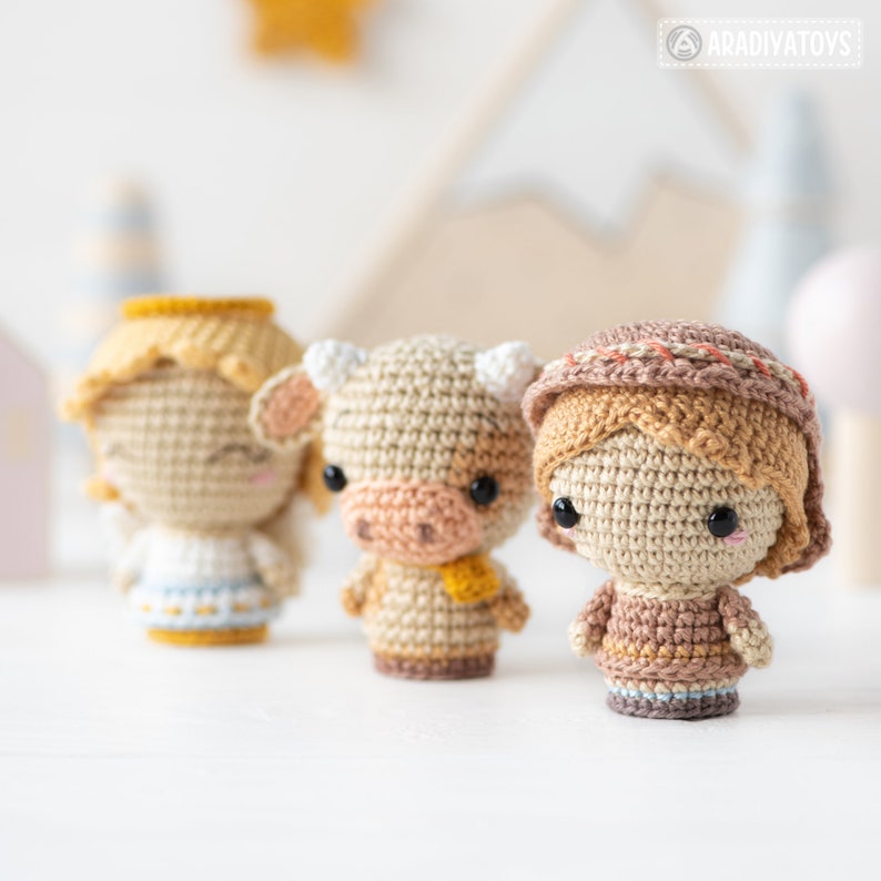 May include: Three crocheted amigurumi toys: an angel, a cow, and a girl wearing a brown hat. The toys are made with beige yarn and have brown eyes. The angel has a halo and a white dress, the cow has a brown nose and a yellow scarf, and the girl has a brown dress and a brown hat with a white trim.