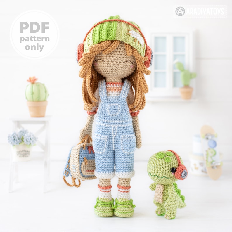 May include: Crocheted doll with brown hair wearing a green and white striped hat, blue overalls, and green shoes. The doll is holding a small green backpack. A small green crocheted dinosaur with headphones is standing next to the doll.