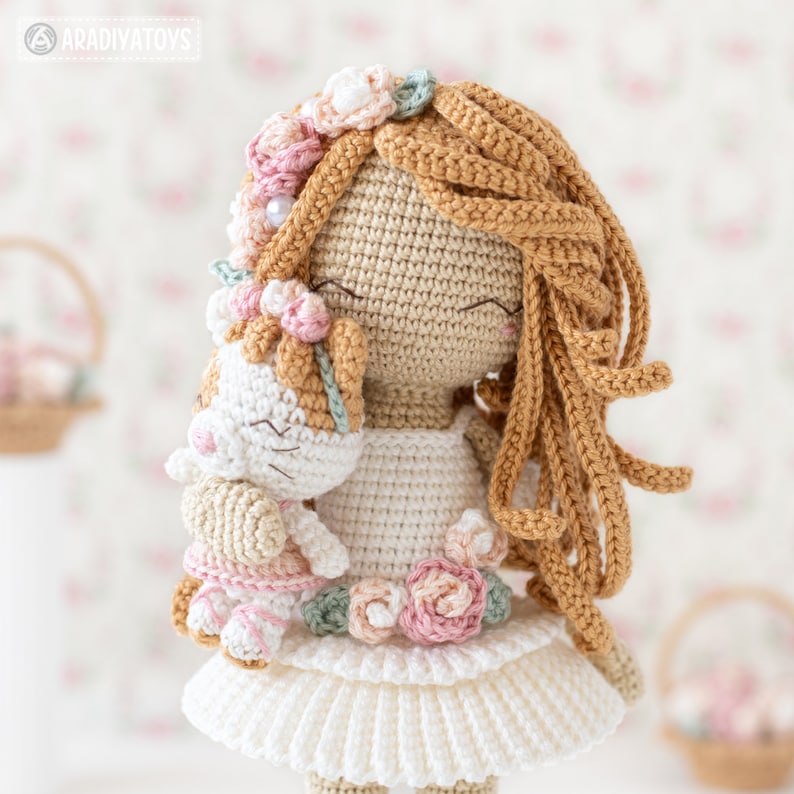 May include: A handmade crochet doll with long, flowing brown hair and a white dress. The doll is holding a smaller crochet animal. The doll's hair is adorned with a floral crown, and a floral embellishment is on the dress. The doll has closed eyes and a gentle expression.