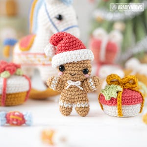 Op de afbeelding: Een gehaakte peperkoekman met een rode en witte kerstmuts. De peperkoekman heeft een witte strik en witte accenten. Andere gehaakte kerstversieringen zijn op de achtergrond te zien.