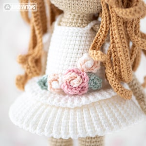 May include: Crocheted doll with long, curly, light brown hair and a white dress. The dress is adorned with a floral embellishment featuring pink, peach, and green crocheted flowers. The doll's details are meticulously crafted.