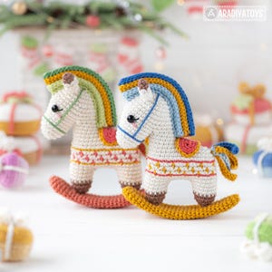 May include: Two crocheted rocking horse toys. The horses are white with colorful manes and saddles. One has a green and yellow mane, the other has a blue and yellow mane. Both have brown rockers and are decorated with red and yellow patterns.