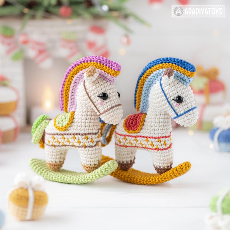 May include: Two crocheted rocking horse toys. The horses are white with colorful manes and tails, and decorative saddles. The rockers are green and yellow. The background includes Christmas decorations.