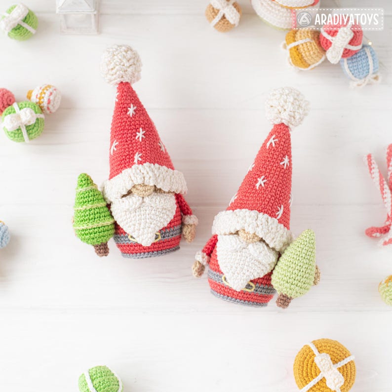 K&ouml;nnte beinhalten: Zwei geh&auml;kelte Weihnachtsmann-Zwerge mit roten M&uuml;tzen und wei&szlig;en B&auml;rten. Jeder Zwerg h&auml;lt einen gr&uuml;nen Weihnachtsbaum. Die M&uuml;tzen haben wei&szlig;e Sternakzente. Andere geh&auml;kelte Weihnachtsschmuckst&uuml;cke sind verstreut.