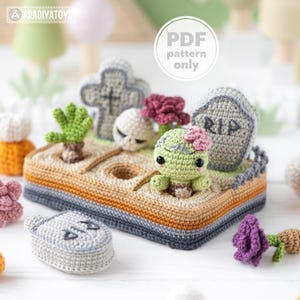 May include: Crocheted graveyard scene with a zombie, tombstones, and flowers. The scene includes a zombie with a pink flower, a skull, and a cross. The base is a rectangle with a gray, orange, and tan border. The text "PDF pattern only" is in a white circle.