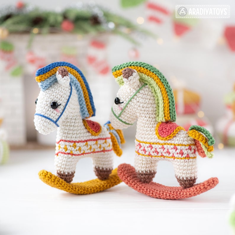 May include: Two handmade crochet rocking horses. The horses are white with colorful manes and saddles. The rocking bases are yellow and coral. The horses have black eyes and are decorated with red, yellow, and blue patterns. The background is blurred.