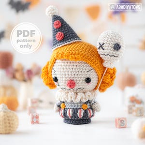 Puede incluir: Una muñeca payaso de ganchillo con pelo naranja, un sombrero cónico gris con detalles rojos y un cuello de volantes. La muñeca viste un traje a rayas grises y blancas con botones naranjas y sostiene un globo blanco con una cara cosida. Texto PDF pattern only.