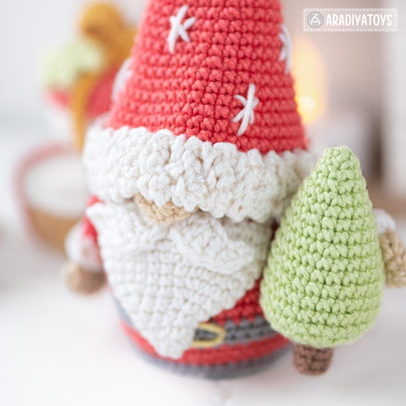 K&ouml;nnte beinhalten: Eine handgefertigte H&auml;kelzwerg-Puppe mit roter M&uuml;tze mit wei&szlig;en Sternen, einem wei&szlig;en Bart und einem gr&uuml;nen Weihnachtsbaum. Der Zwerg tr&auml;gt ein rot-graues Outfit mit einem goldfarbenen G&uuml;rtel. Die Puppe ist eine festliche Weihnachtsdekoration.