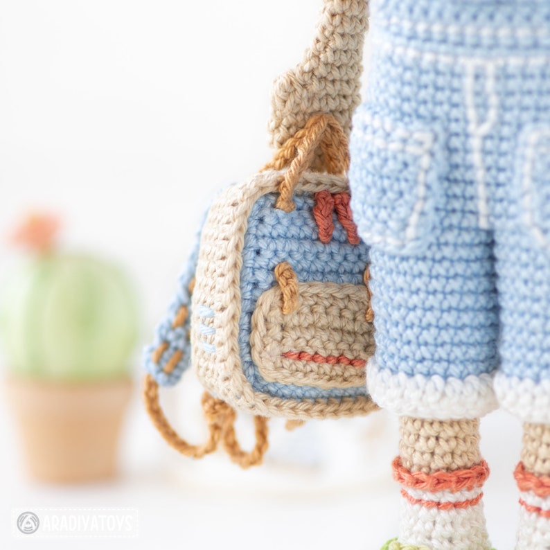 May include: A crocheted doll wearing blue overalls and a beige backpack with orange straps. The doll is wearing white socks with orange stripes.