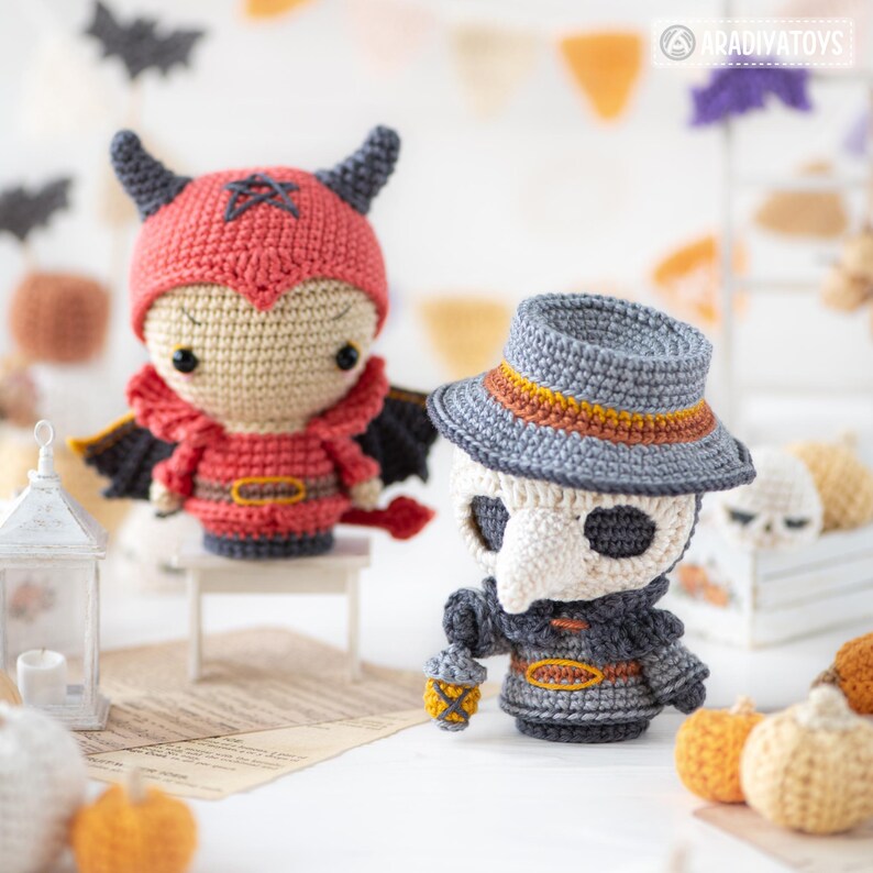 May include: Two crocheted dolls: one red devil figure with horns, wings, and a pentagram on its hood, and one gray plague doctor figure wearing a wide-brimmed hat, a long beak mask, and holding a small lantern, both featuring detailed yarn stitching.