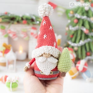 K&ouml;nnte beinhalten: Handgefertigte geh&auml;kelte Weihnachtsmann-Figur mit roter M&uuml;tze mit wei&szlig;en Schneeflocken und einem wei&szlig;en Bommel. Der Weihnachtsmann h&auml;lt einen kleinen gr&uuml;nen Weihnachtsbaum. Die Figur hat einen wei&szlig;en Bart, einen grauen G&uuml;rtel und einen roten Anzug.
