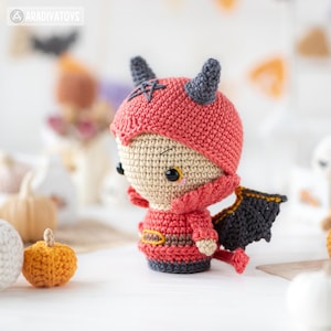 May include: A crocheted amigurumi doll featuring a red hooded outfit with dark gray horns and a black pentagram symbol on the forehead. The doll has black wings, a small red tail, and a tan face with black eyes. A small orange crocheted pumpkin sits nearby.