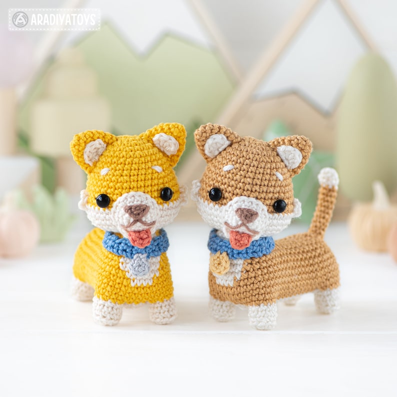 K&ouml;nnte beinhalten: Zwei geh&auml;kelte Shiba Inu-Pl&uuml;schtiere, eines gelb und eines braun, mit wei&szlig;en Akzenten, blauen Halsb&auml;ndern und rosa Zungen. Die Pl&uuml;schtiere stehen auf einer wei&szlig;en Oberfl&auml;che.