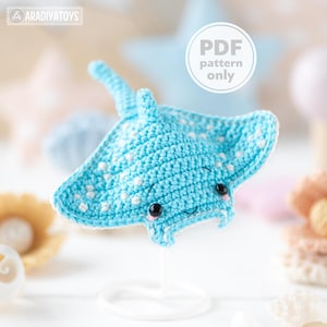 Könnte beinhalten: Gehäkelter blauer Mantarochen mit weißen Punkten und schwarzen Augen. Das handgefertigte Spielzeug hat ein lächelndes Gesicht und ist auf einem weißen Ständer montiert. Der Text "PDF pattern only" ist enthalten.
