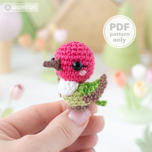 Peut inclure: Un petit colibri en crochet avec une tête rose vif, une poitrine blanche et des ailes vert clair aux bords marron. L'oiseau possède des yeux en perles noires et un bec marron. Un graphique circulaire blanc avec le texte PDF pattern only est visible.