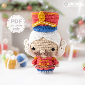 May include: A crocheted nutcracker doll in a red and gold uniform, with white hair and a mustache. The doll has a red and blue hat with a gold accent. The text "PDF pattern only" is in the upper left corner. A festive holiday decoration.