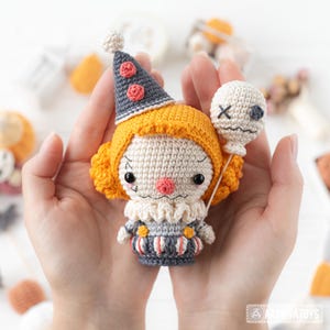 May include: A crocheted clown doll featuring orange hair, a grey cone hat with red accents, a ruffled white collar, and a grey striped outfit. The doll holds a small white balloon with a stitched face and has black eyes and a red nose.