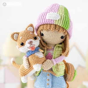 K&ouml;nnte beinhalten: Eine geh&auml;kelte Puppe mit einem rosa-gr&uuml;nen Hut, einer rosa-gr&uuml;nen Weste und einer blauen Jeans. Die Puppe h&auml;lt einen geh&auml;kelten braunen und wei&szlig;en Hund.