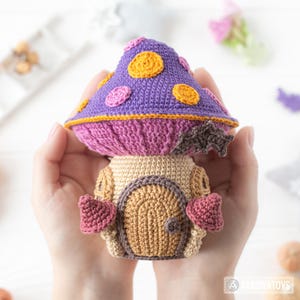 May include: A handmade crochet mushroom house with a purple cap adorned with orange and pink circles. The mushroom has a tan base with a brown door and pink heart-shaped accents. The item is held in two hands.