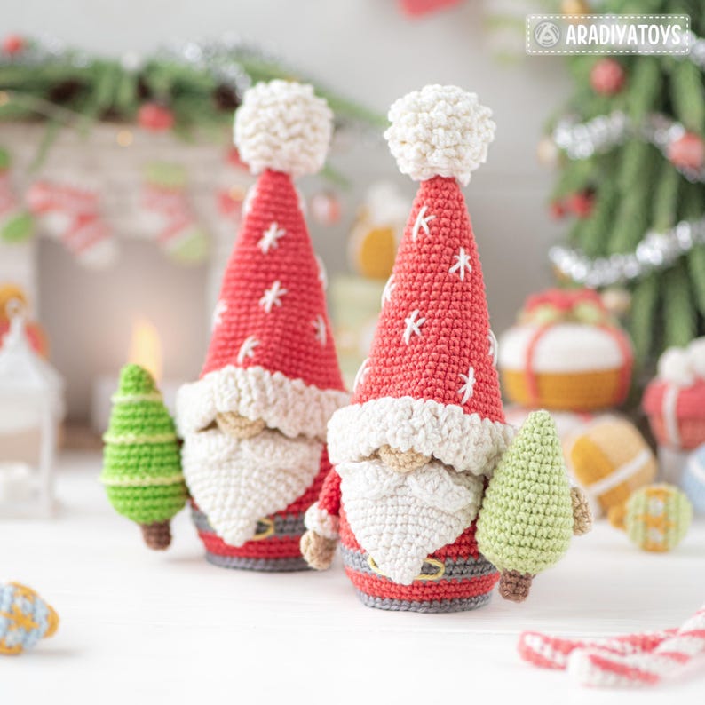 K&ouml;nnte beinhalten: Zwei handgefertigte geh&auml;kelte Weihnachtszwerge mit roten M&uuml;tzen und wei&szlig;en B&auml;rten. Die M&uuml;tzen haben wei&szlig;e Schneeflocken-Akzente und einen wei&szlig;en Bommel. Neben jedem Zwerg befindet sich ein kleiner gr&uuml;ner geh&auml;kelter Weihnachtsbaum. Der Hintergrund enth&auml;lt Weihnachtsdekorationen.