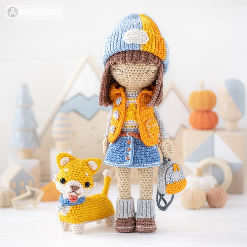 K&ouml;nnte beinhalten: Eine geh&auml;kelte Puppe mit einem blauen und orangenen Hut, einer gelben Weste, einem blauen Jeansrock und grauen Stiefeln. Die Puppe h&auml;lt einen kleinen blauen und orangenen Rucksack. Ein geh&auml;kelter gelber Hund mit roter Zunge steht neben der Puppe.