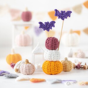May include: A stack of three crocheted pumpkins in orange, white, and burgundy, topped with two purple bat decorations on wooden sticks. Other crocheted pumpkins in various colors and sizes are in the background, along with decorative bunting.