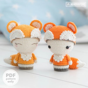 May include: Two crocheted fox amigurumi toys with orange and white fur, wearing white crocheted hats. The toys are sitting on a white wooden surface. PDF pattern only.