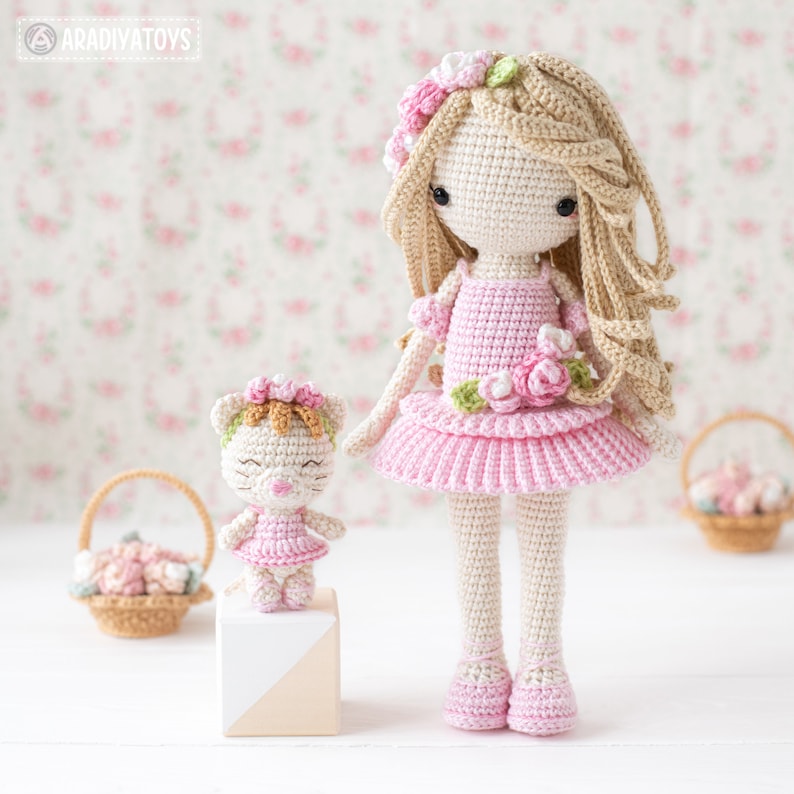May include: Two handmade crochet dolls. One is a girl doll with long blonde hair, wearing a pink dress with floral accents. The other is a small cat doll, also in a pink dress. Both dolls have floral headbands. The background features a floral pattern.