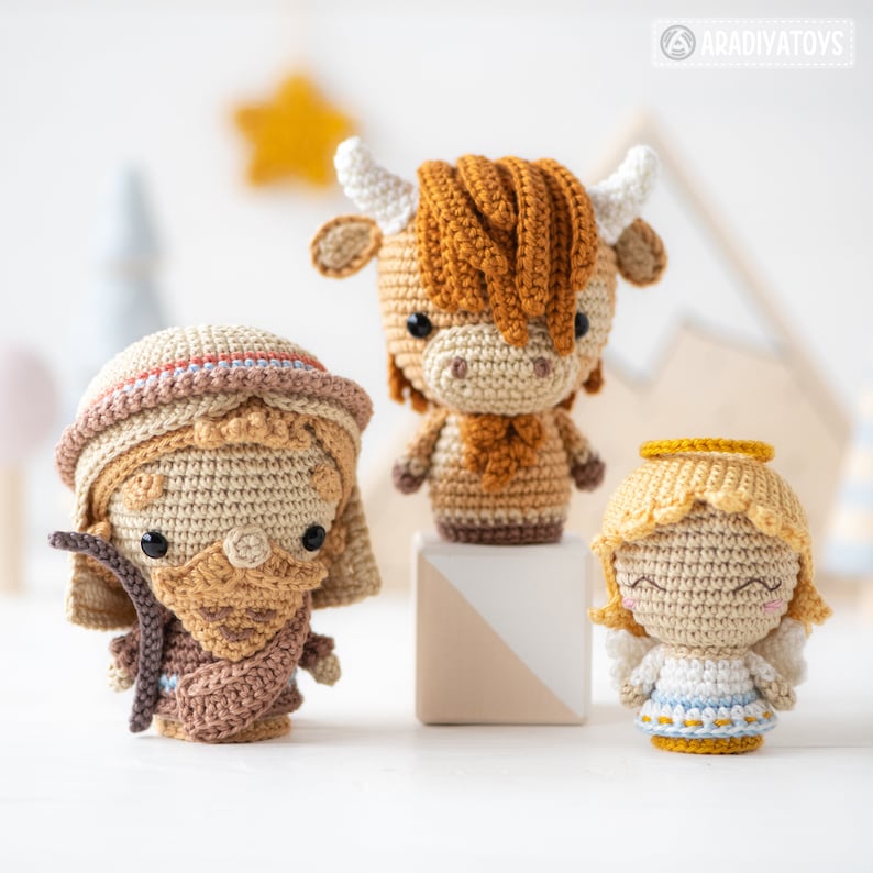 May include: A set of three crocheted amigurumi toys: a brown and white cow, a brown and beige shepherd, and a yellow angel with a blue dress. The toys are arranged on a white surface.