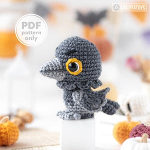 Peut inclure: Un oiseau gris crocheté avec un œil cerclé de jaune et une pupille noire. L'oiseau a un cou à volants et se tient sur un bloc blanc. L'image comprend le texte "PDF pattern only".