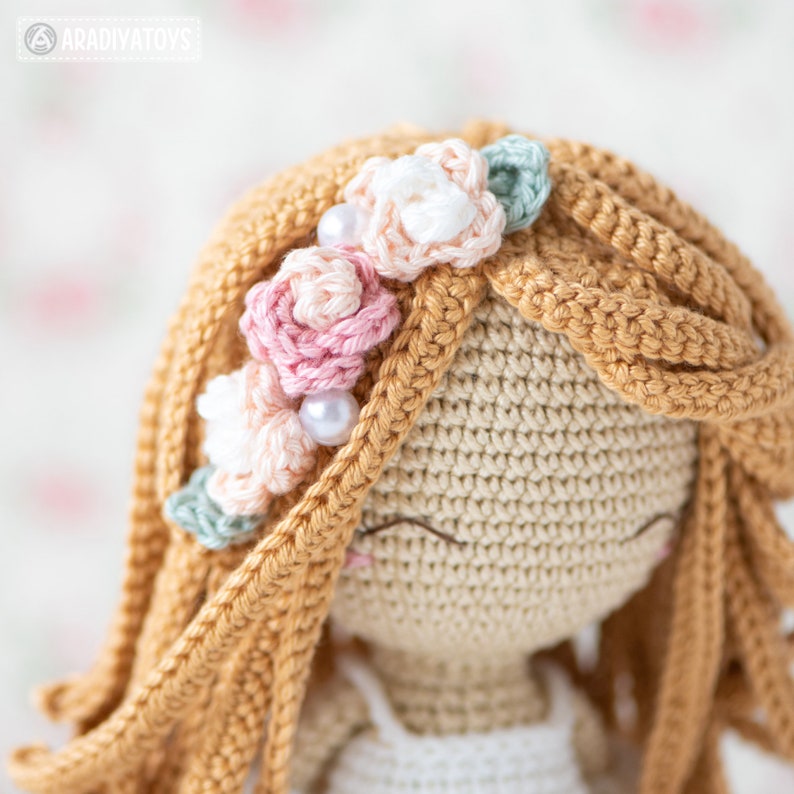 May include: Close-up of a crocheted doll with long, light brown hair and a floral headband. The headband features pink, white, and light green flowers with pearl accents. The doll's face has closed eyes and a small, smiling mouth.