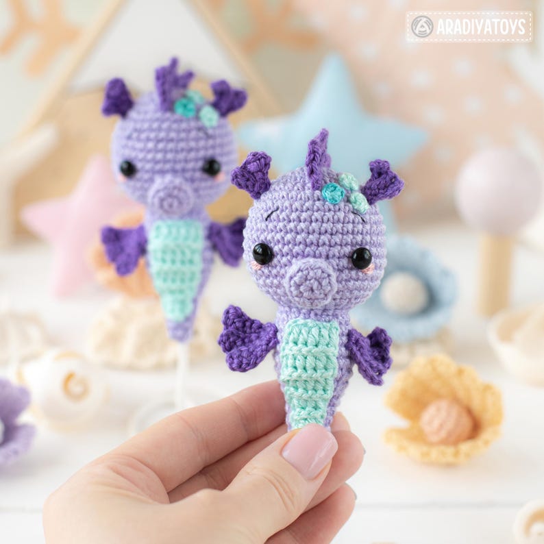 May include: Two handmade crochet seahorse toys. The seahorses are purple with light blue and purple accents. The seahorses have black eyes and are held by a person. The background includes other crochet sea-themed items.