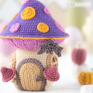 May include: A handmade crochet mushroom house toy with a purple cap adorned with orange and pink circles. The house features a tan body with a brown door and pink heart-shaped accents. A gray bat is attached to the front.