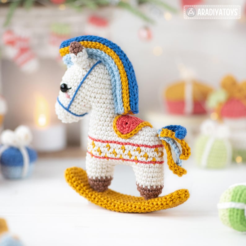May include: A handcrafted crochet rocking horse toy. The toy features a white body with blue, yellow, and red accents. The rocking horse has a yellow base and is decorated with a detailed pattern. The toy is designed for children.